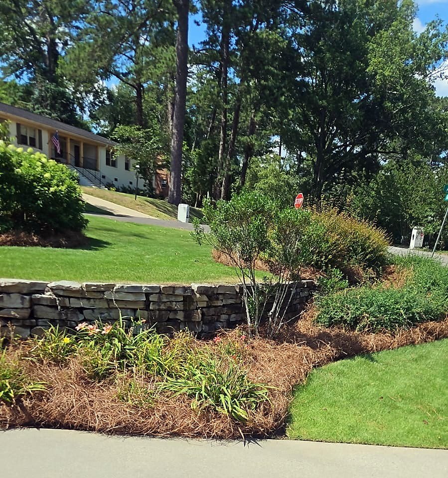 Dry-stack stone retaining wall with daylilies and shrubs in a pine straw bed beside a driveway