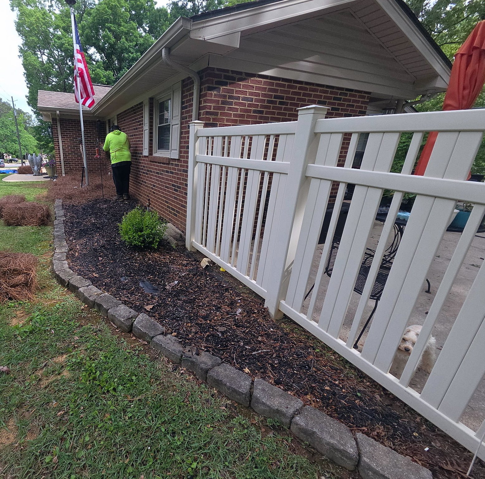Garcia crew member spreading pine straw in a bed by a white fence with bales on the lawn